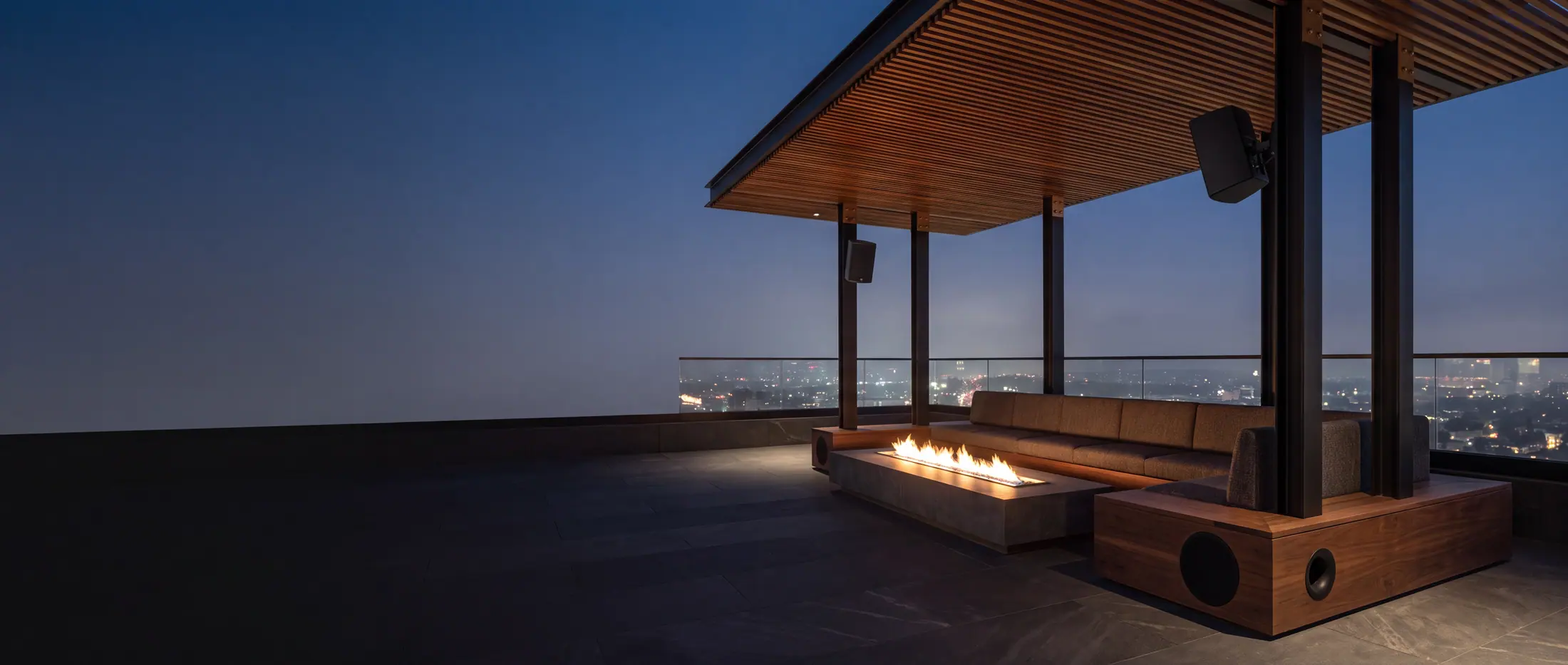 A Los Angeles rooftop terrace at blue hour. Compact loudspeakers are mounted on the pergola columns and a subwoofer is built into the bench plinth, with the city lights below.